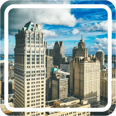 A panoramic view of the Detroit skyline at midday. The sky is a clear blue, and the sun glints off the windows of the skyscrapers.