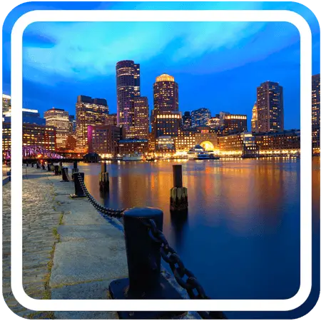 A dock juts out into a calm harbor at night. The city skyline in the background is illuminated by a warm glow