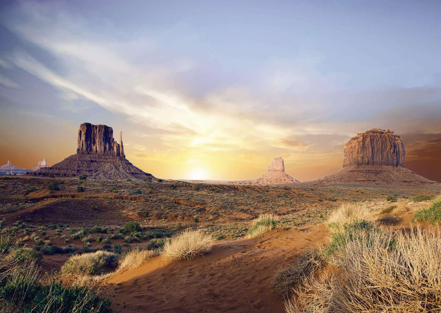 A desert landscape in Monument Valley at sunset. The buttes in the background are silhouettes against a colorful sky.