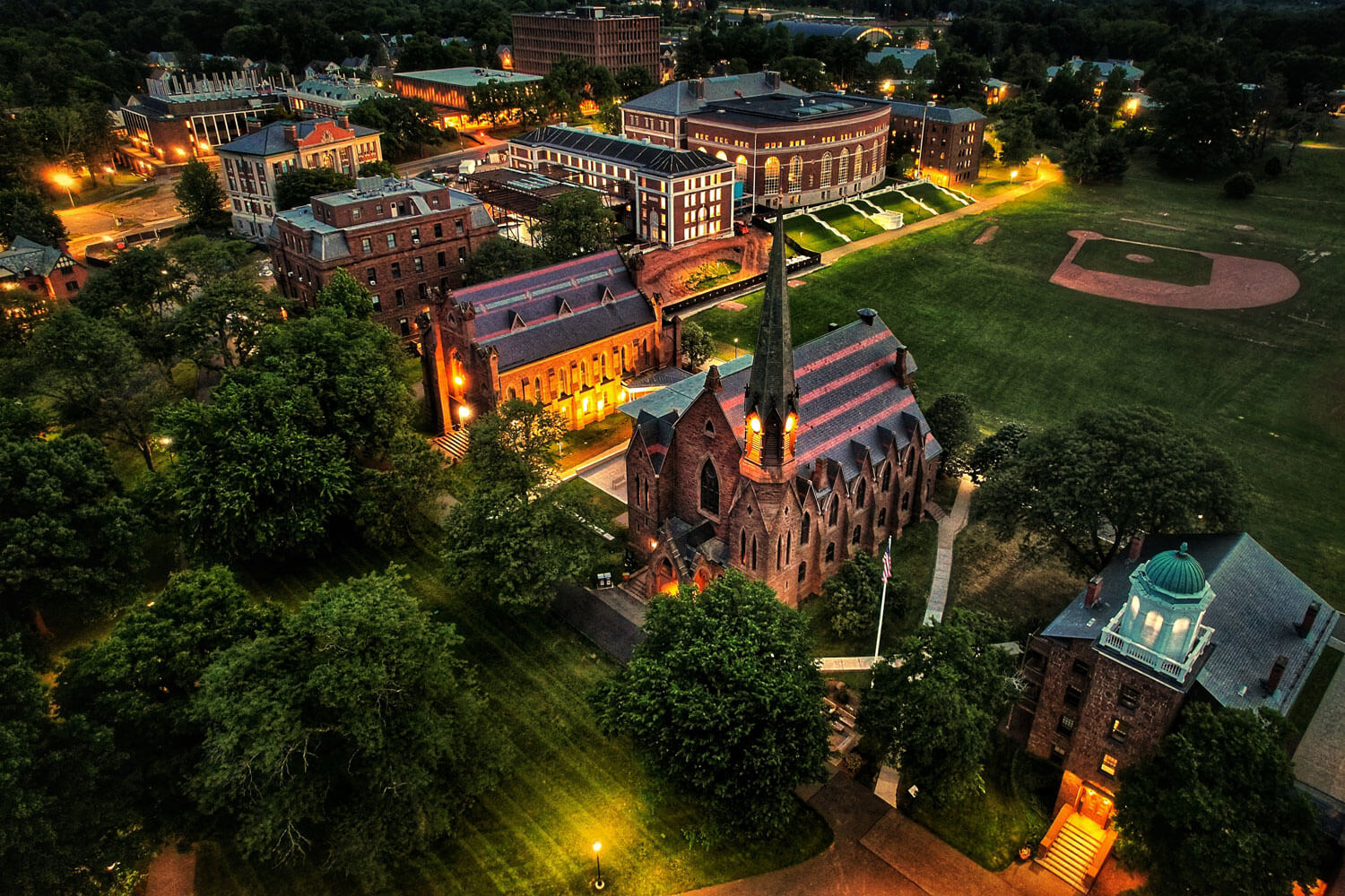 An aerial view of the Wesleyan University campus in Middletown, Connecticut, at night. The buildings are illuminated, with walkways and trees visible throughout the grounds
